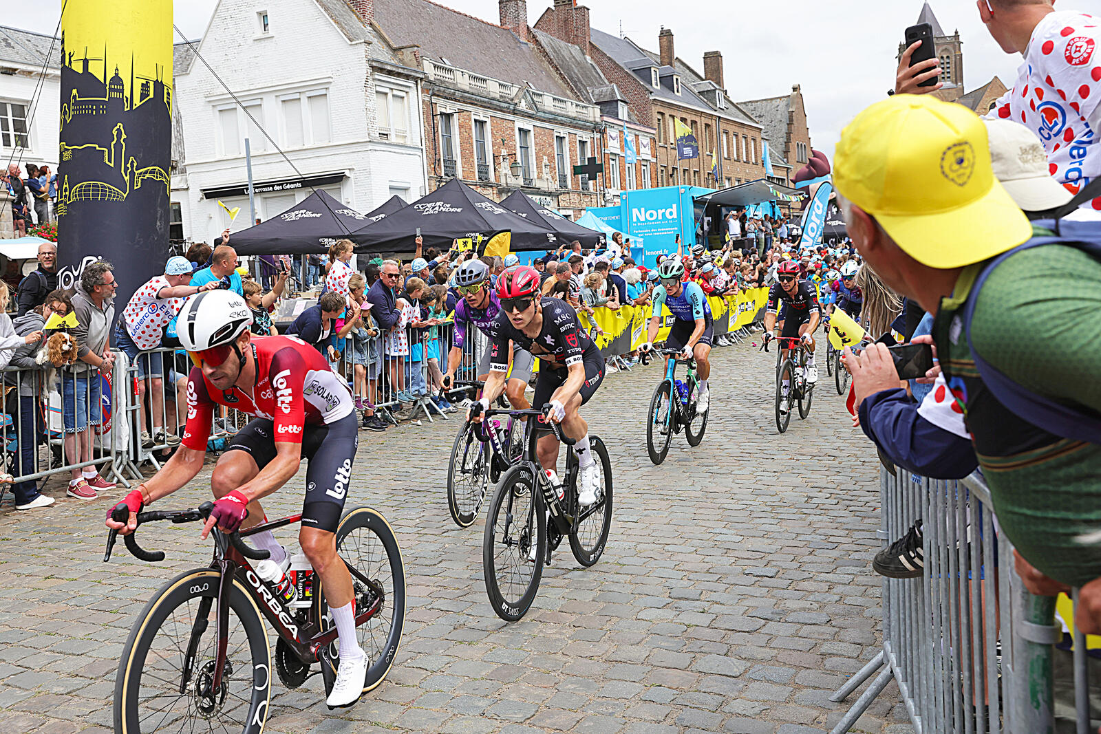 L'image montre des coureurs cyclistes sur la Grand'Place de Cassel, et la foule amassée le long des barrières