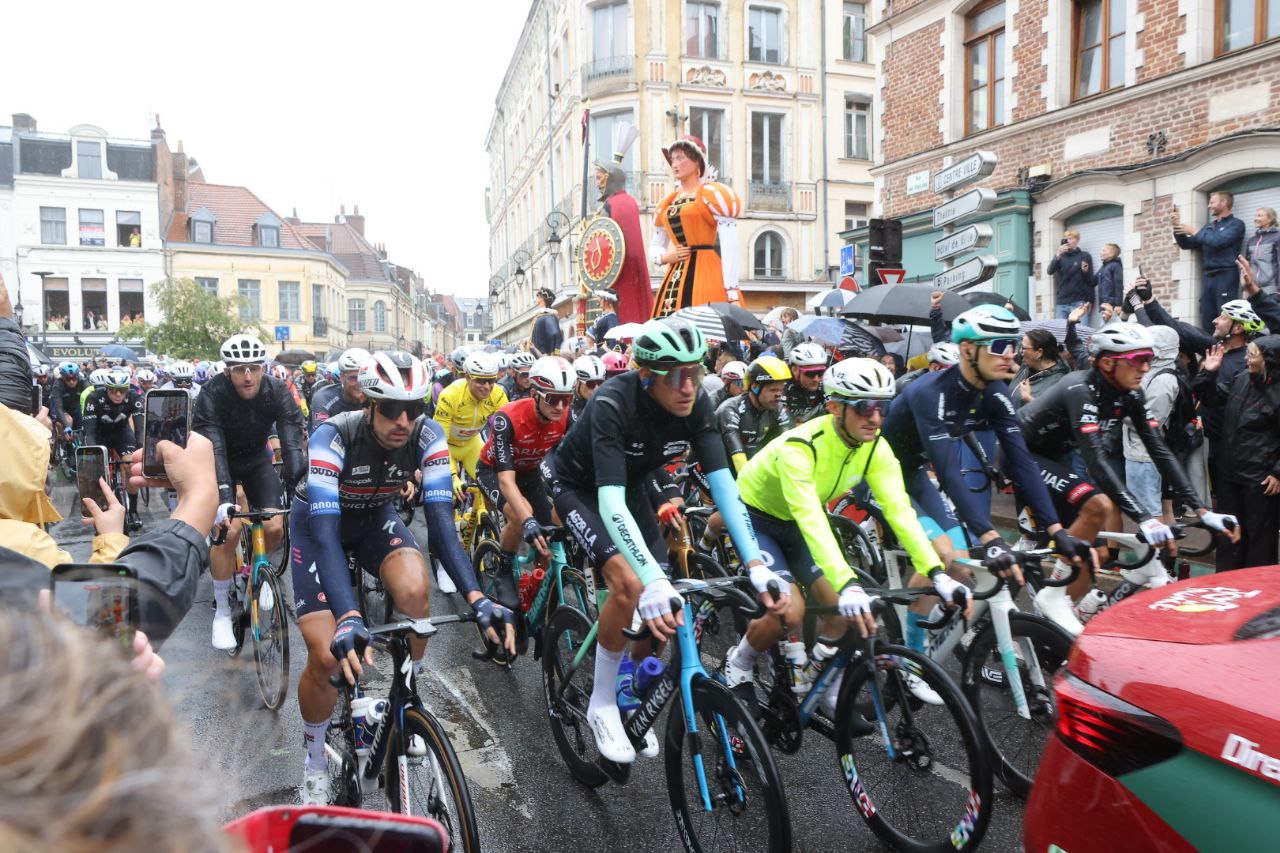 L'image montre des coureurs cyclistes au premier plan, et les géants Monsieur et Madame Gayant en arrière plan.